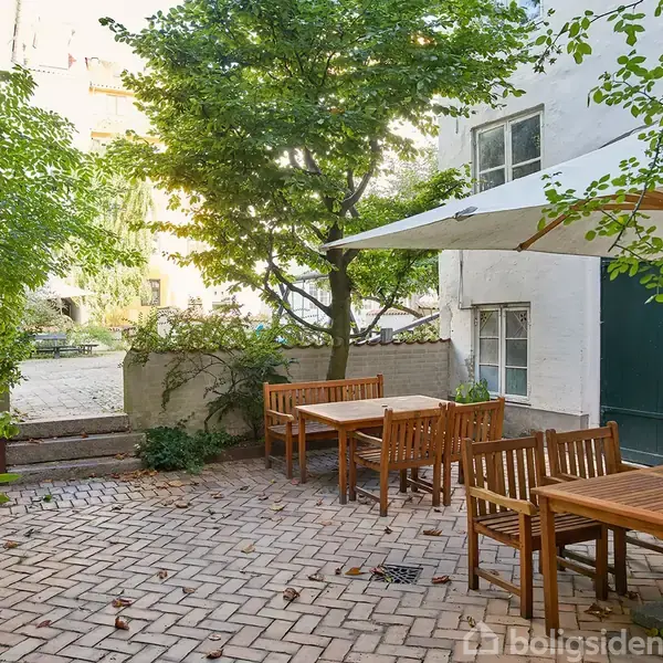 Træmøbler under en parasol står på en brostensbelagt terrasse. Grønt løv omkranser området, og en hvid bygning med vinduer danner baggrund. Ingen tekst er til stede.