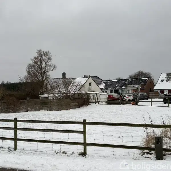 Et snedækket landskab med en indhegnet mark i forgrunden. Flere huse med skråtag i baggrunden, hvoraf nogle har solpaneler. Biler er parkeret ved siden af et af husene.