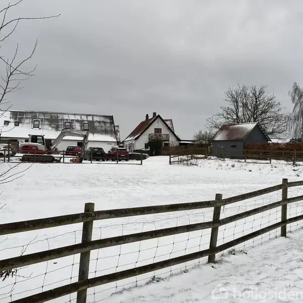 En gård dækket af sne med flere bygninger og biler parkeret foran. Et hegn løber langs en snedækket mark i et vinterlandskab.