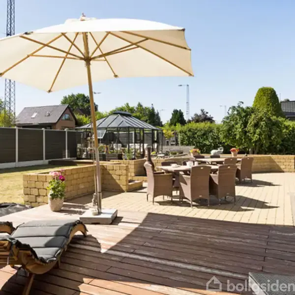 En terrasse med liggestol og bord-sæt under en stor parasol. Omkring er en have med grønne buske og et drivhus. Solen skinner klart på området.