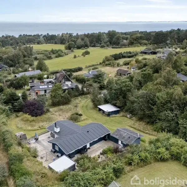 Et sort, lavt hus omgivet af græsplæne og træer ligger blandt andre huse i et frodigt, landligt område nær kysten, med havet synligt i horisonten.