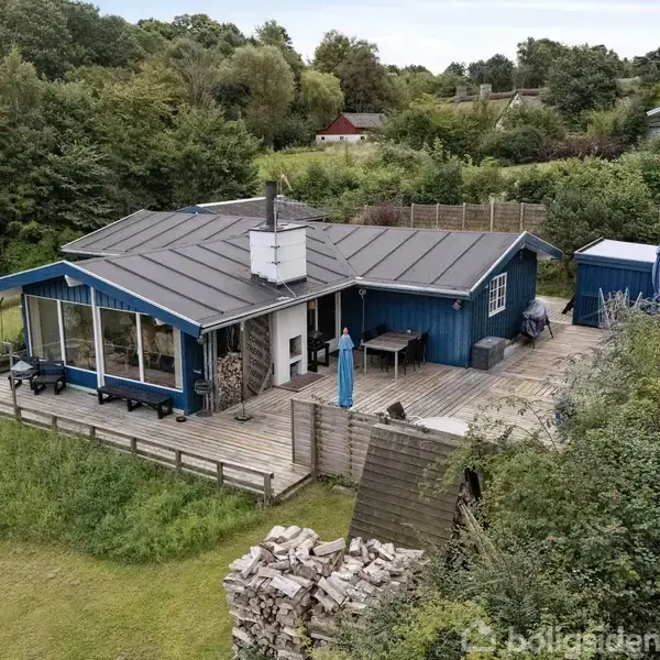 Et blåmalet sommerhus står stille på en grøn græsplæne omkranset af frodige buske. Et stort træterrasseområde omringer huset, hvor en parasol er opslået.