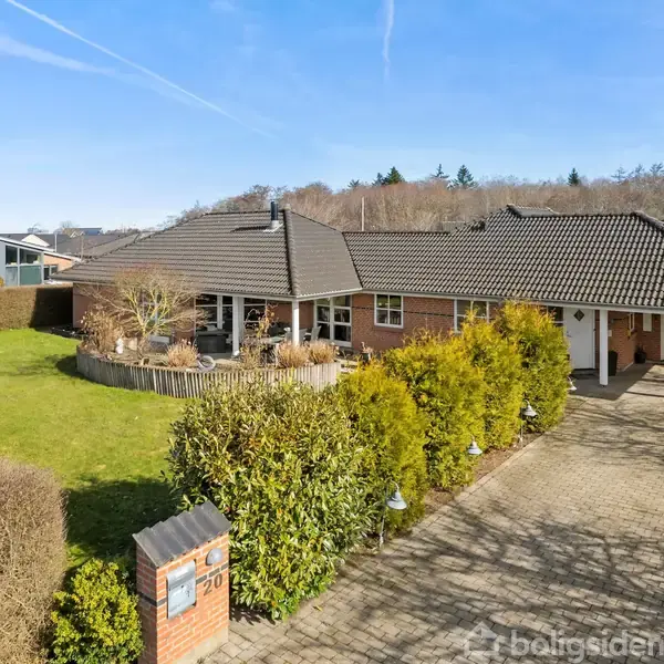 Et étplanshus med mørkt tegltag står på en velplejet grund med grøn græsplæne og hække. En flisebelagt indkørsel fører til en carport. Skyfri blå himmel i baggrunden.