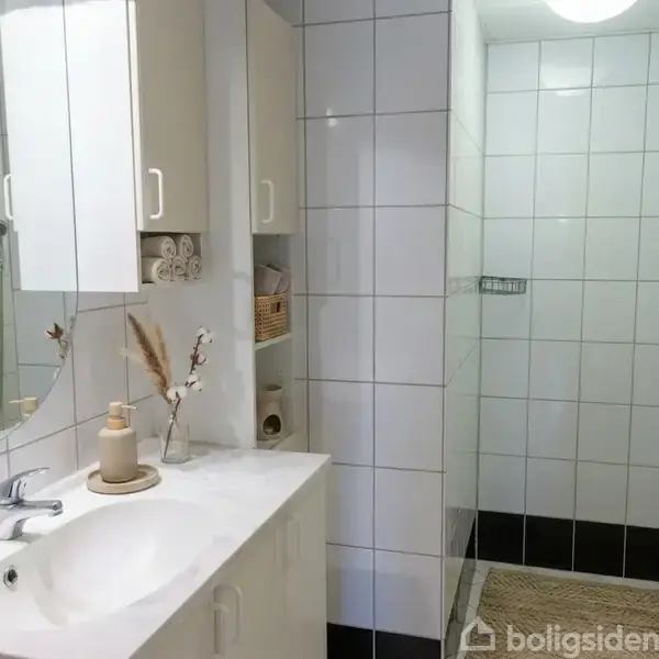 Hvid klinkebelagt badeværelse med håndvask og spejl. Hylder med sammenrullede håndklæder og dekorationer til højre. Bagved indgang til brusekabine med bruser og glasvægge.