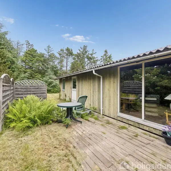 Et træhus med store vinduer vender mod en træterrasse. På terrassen står et grønt bord med stole. Omgivet af buske og træer i en frodig have.