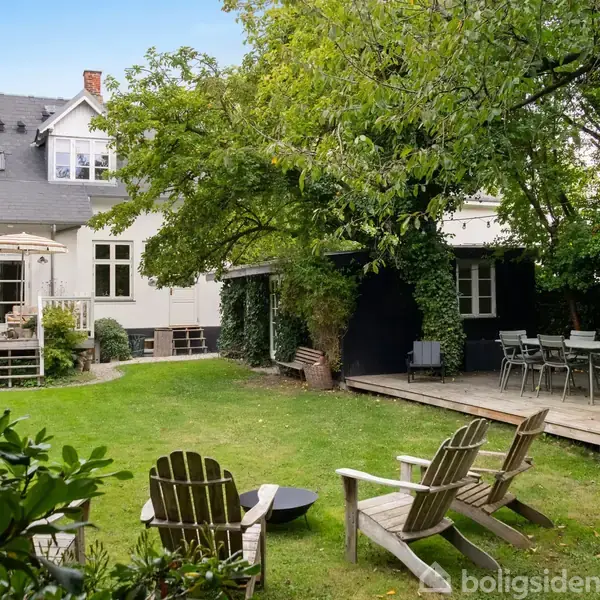 En hvid villa med sort tag står midt i en frodig have. Træstole er placeret omkring en terrasse, mens havemøbler står på en træplatform omgivet af grønne træer og buske.