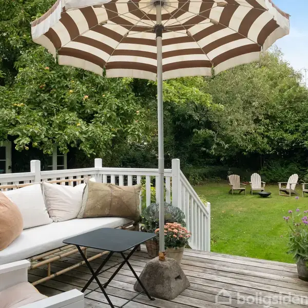 Terrasse med hvid bænk og puder, under stribet parasol. Lille sort bord ved siden af planter i krukker. I baggrunden ses græsplæne med stole samt tætte grønne buske og træer.