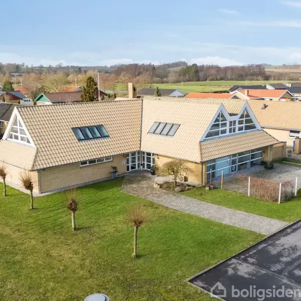 Hus med skråtag og store vinduer, omgivet af velplejede græsplæner og træer, ligger i et villakvarter med flere huse og biler parkeret ved siden af asfalteret vej under en blå himmel.