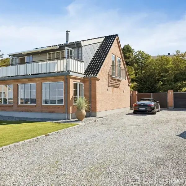 Et to-etagers, gult murstenshus med stor altan står på en grusbelagt indkørsel. En bil er parkeret ved siden af huset, omgivet af græsplæne og træer i baggrunden.