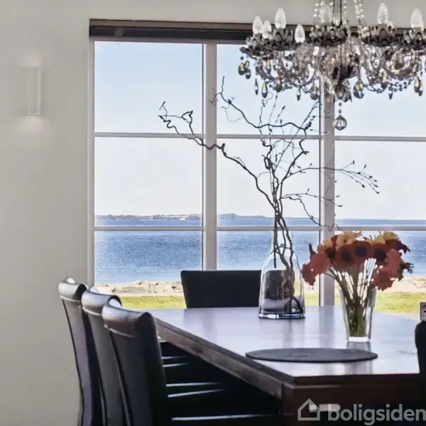 Et spisebord med blomster og grene i vaser står i en lys spisestue. En lysekrone hænger over bordet. Udsigt til havet kan ses gennem de store vinduer.