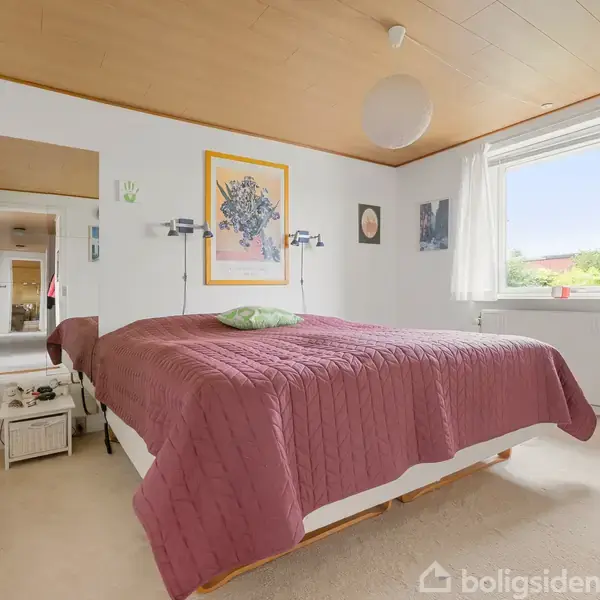 Seng med en lyserød sengetæppe står i et lyst soveværelse. Væggene er dekoreret med kunst, og et stort vindue slipper naturligt lys ind. En lampe hænger fra loftet.