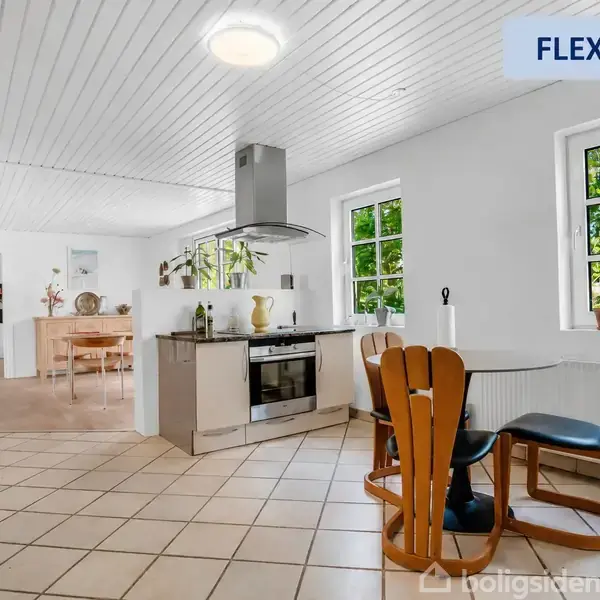 Et moderne køkken-alrum med træmøbler og flisegulv. Et spisebord med stole står ved siden af vinduer med udsigt til grønne træer. Øverst til højre står teksten "FLEXBOLIG".