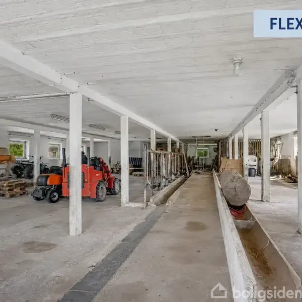 En traktor står stille i et rummeligt, hvidmalet lager med betongulv og søjler. Et skilt øverst siger "FLEXBOLIG". Værktøj og materialer ligger langs væggene.