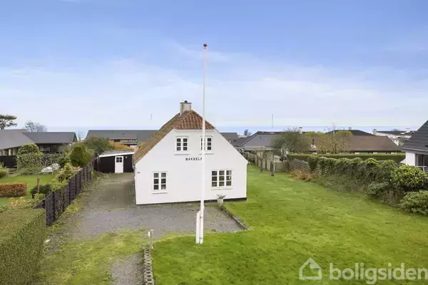 Hvidt hus med skilt "BAKKELEJ" står på en grøn græsplæne med flisegang. Bagved er et skur, og haver omgiver ejendommen i et kystnært boligområde med udsigt til havet.