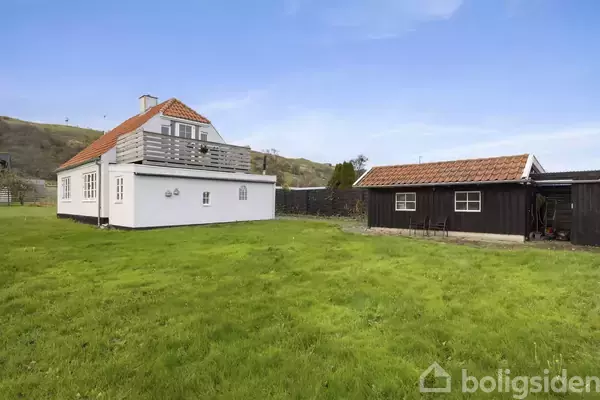 Et hvidt hus med en tagterrasse står i en grøn have ved siden af en sort trægarage. Omkring huset ses bakket landskab og blå himmel.