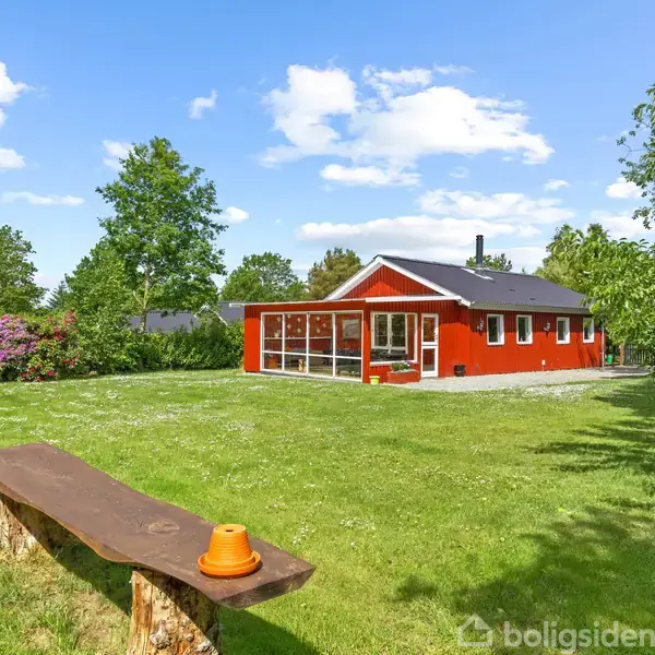 Rødt hus står stille i en frodig have omringet af grønne træer. En træbænk med orange blomsterpotte er placeret på græsset, mens himlen er blå med hvide skyer.