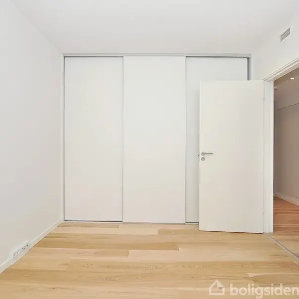 En tomt rum med indbygget hvid skydedørsgarderobe, åben hvid dør til højre og lyst trægulv. Væggene er hvide og der er ingen synlige møbler til stede.
