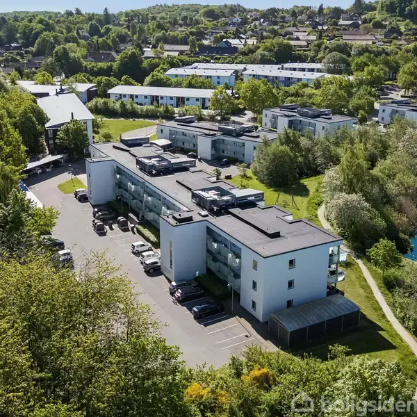 Lejlighedskompleks set ovenfra omgivet af grønne træer og buske, parkerede biler foran bygningen. I baggrunden ses flere bygninger og en blå sø til højre.