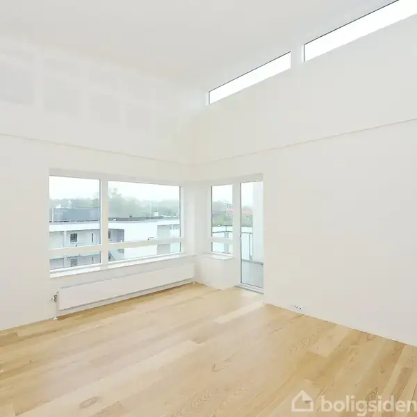 Et lyst rum med trægulv og store vinduer giver udsigt til en balkon og bygninger udenfor. Højt til loftet og minimalistisk indretning.