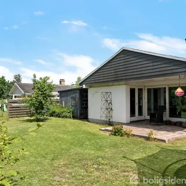 Hus set fra haven med åben terrasse, havemøbler, græsplæne og omkransende hække. Til højre en trampolin. Skyfri himmel skaber en lys og solrig dag.