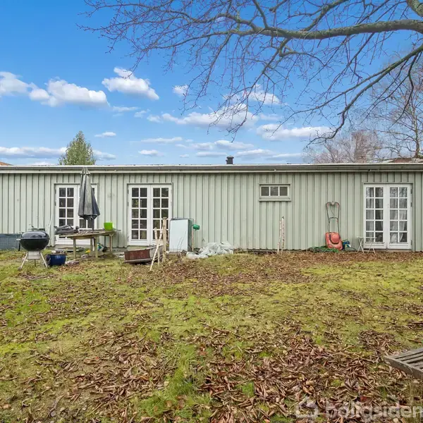 En enkel, lav bygning med to døre og et vindue. Den står i en have omgivet af træer og græs. Bord og grill er placeret udenfor.