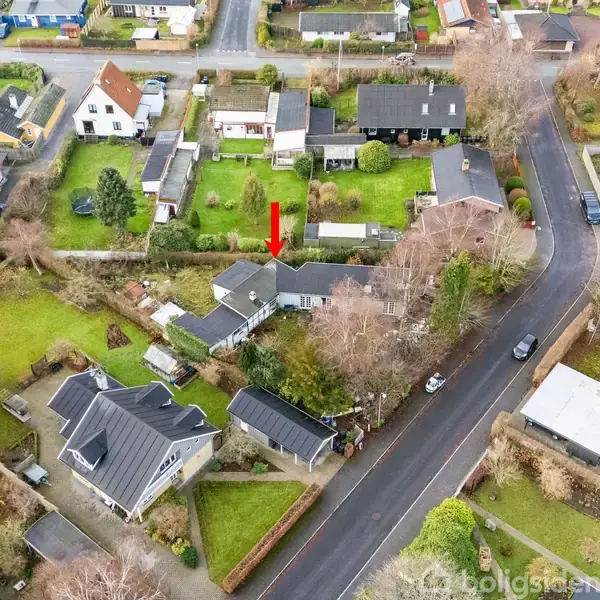 En rød pil peger ned mod et hus omgivet af træer og haver. En vej løber forbi, hvor biler er parkeret, i en beboelseskvarter med flere huse og grønne områder.