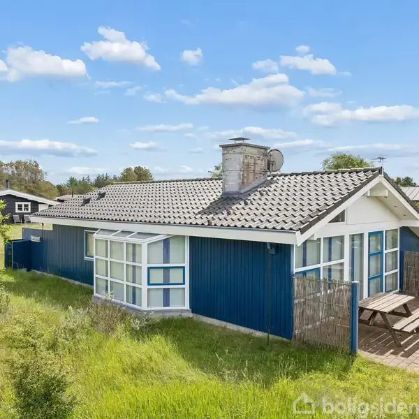 Blåt sommerhus med gråt tag står på græsareal. Huset har store vinduer og en udendørs terrasse med bord og bænke. Omgivet af grønne marker og et klart, skyet himmel.