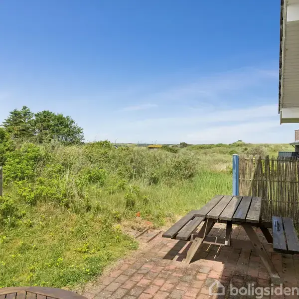 En træbænk med bord står på en terrasse af mursten. Omkring er der en frodig have med buske og græs, under en klar blå himmel.