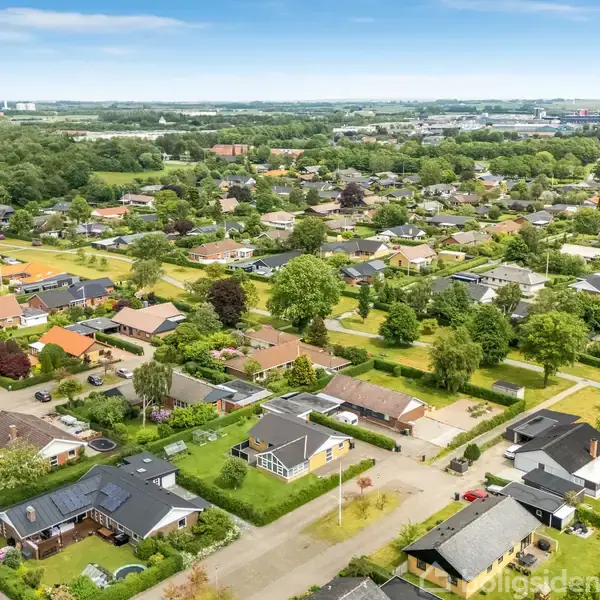 Boligområde med huse spredt ud over et grønt landskab. Huse er omgivet af grønne haver og træer. Baggrunden viser yderligere bebyggelse og skovområder i horisonten.