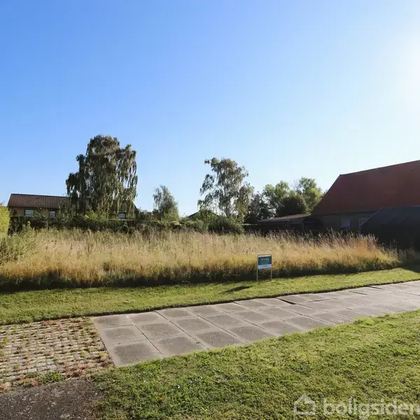 Græsplæne med højt græs vokser foran huse. En lille skilt står midt på grunden. Omkringliggende huse og træer skaber en fredelig, solrig atmosfære.