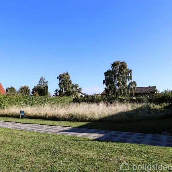 En græsplæne med en flisebelagt sti løber gennem en velplejet have. Høje træer grænser til området, og der er huse i baggrunden under en skyfri blå himmel.