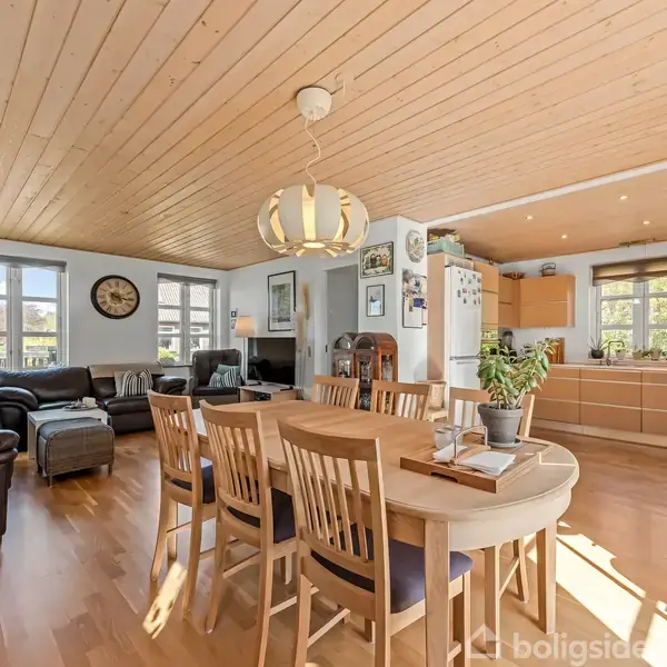 Spisebord med seks stole står i lys, åben stue-køkken. Stuen har brun lædersofa, vægure, og indendørs planter. Lyst træloft og gulv skaber en varm atmosfære.