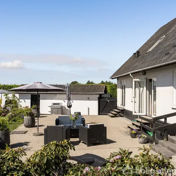 Hus med hvid facade og sort tag står i sollys. Foran er en terrasse med sorte havemøbler og parasol. Haven har grønne planter, og der ses åbne marker i baggrunden.