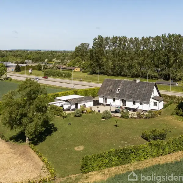 En hvid bygning med sort tag står i en grøn, åben have. Vejen i baggrunden er omgivet af marker og træer under en klar blå himmel.