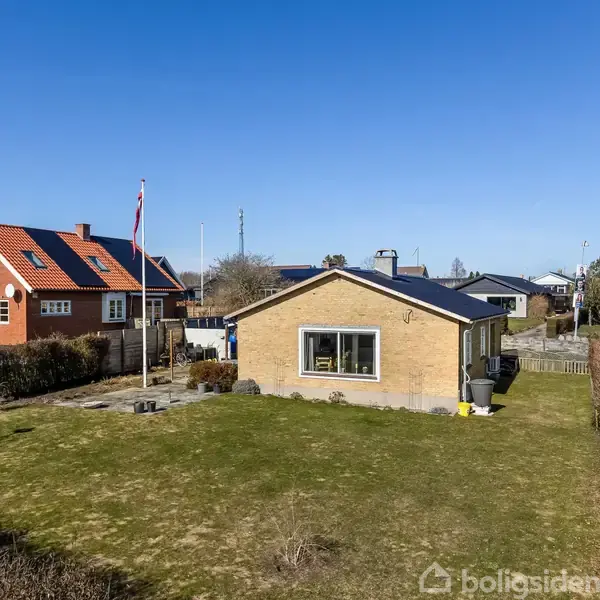 Et gult murstenshus står på en græsplæne med en flagstang i nærheden. Baggrunden har blå himmel og flere huse i et boligområde.