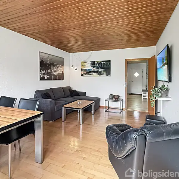 Stue med grå sofa og lænestol centreret omkring et sofabord. Et spisebord med sorte stole står til venstre. Der er moderne kunst på væggene og træloft.