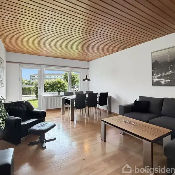Stue med sort sofa og lænestol, sofabord og spisebord med seks stole. Store vinduer giver udsigt til grønne omgivelser. Lyse trægulve og træloft skaber en varm atmosfære.