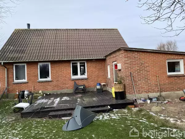 En rød murstenshus med skråtag, tre vinduer, og en træterrasse i baghaven. Diverse genstande og en parabol ligger på græsset, og der er lidt sne på jorden.