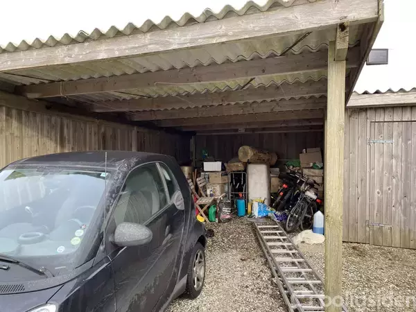En sort bil holder parkeret i en carport. Til venstre ses en stige og diverse genstande samt træ og en cykel. Miljøet er præget af et grusunderlag og trævægge.