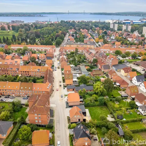 Gader med røde tage strækker sig gennem en forstad med træer og haver. Bebyggelsen omgiver et grønt område. I baggrunden ses vand og en bro.