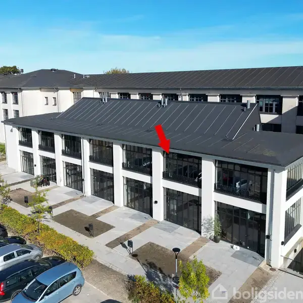 Bygning med moderne facade og store vinduer, pil peger på bestemt sektion. Omkringliggende område med parkeringsplads og biler, græs og blå himmel i baggrunden.