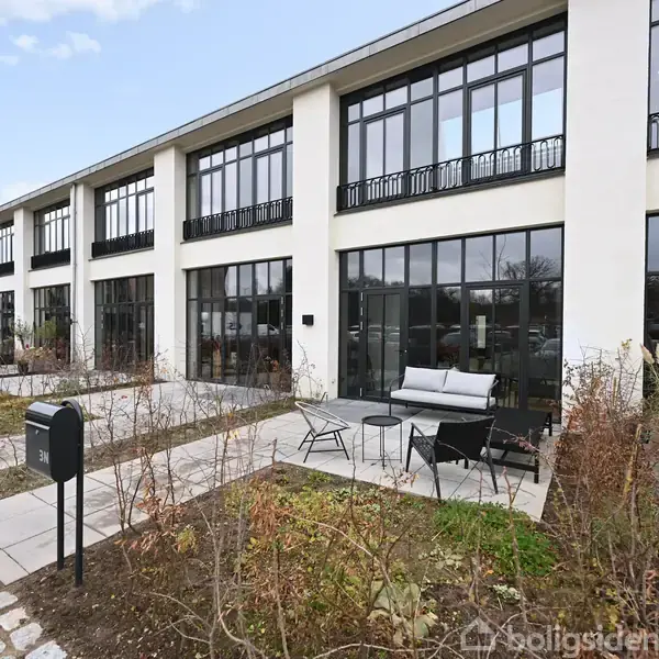 Køkkenet har grå skabe og en hvid bordplade, integreret vask og kogeplade. Store vinduer giver udsigt til en udendørs parkeringsplads omgivet af grønne buske.