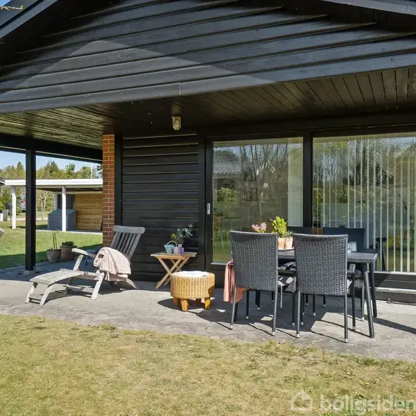 Sort træhus med overdækket terrasse. Terrassen har flisebelægning og indeholder hængekøjestol, liggestol, bord med fire stole og blomsterkrukker. Græsplæne omgiver terrassen, og træer ses i baggrunden.