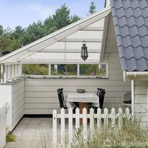 Lille, overdækket terrasse med hvidt træhegn, to sorte stole ved et bord. Græsområde og andre huse i baggrunden. Nummer "10" på husvæggen.