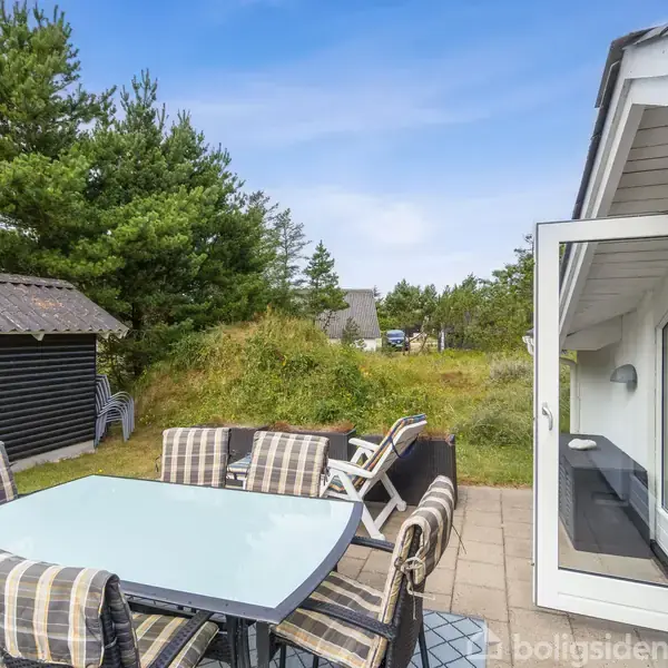Terrassebord med glasplade og seks stole står på en flisebelagt terrasse. Døren til et hus står åben. Omgivet af grønne træer og et mindre skur i baggrunden.