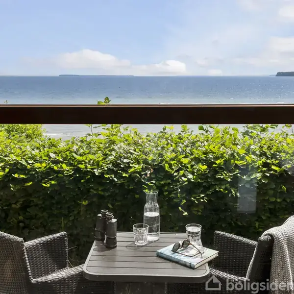 To stole og et bord med kikkert, glas og en karafel står på en terrasse med udsigt over en busk og havet. Himlen er blå med få skyer.