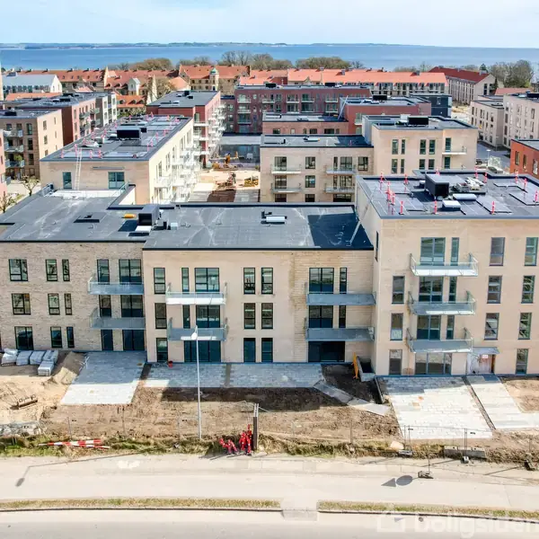 Et moderne lejlighedskompleks under opførelse med flere etager, stor facade med altaner, omgivet af en byggeplads. I baggrunden ses en by og en fjern vandudsigt.