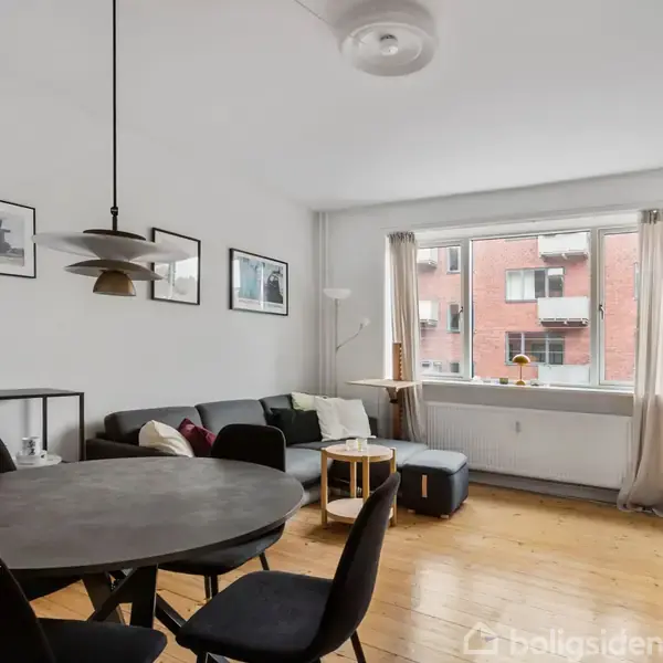 Stue med rundt spisebord og stole, sofa nær et vindue, og væg dekoreret med billeder. Udenfor ses en rød murstensbygning. Rummelig, lyst indrettet med træmøbler.