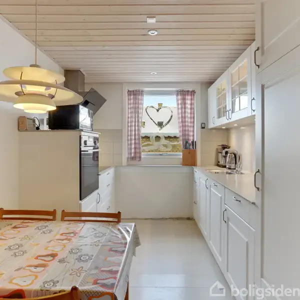 Køkkenbord dækket med voksdug og stole ved siden af moderne ovn. Over bordet hænger en lampe. I baggrunden ses et vindue med lyserøde gardiner og hjerteformet dekoration.