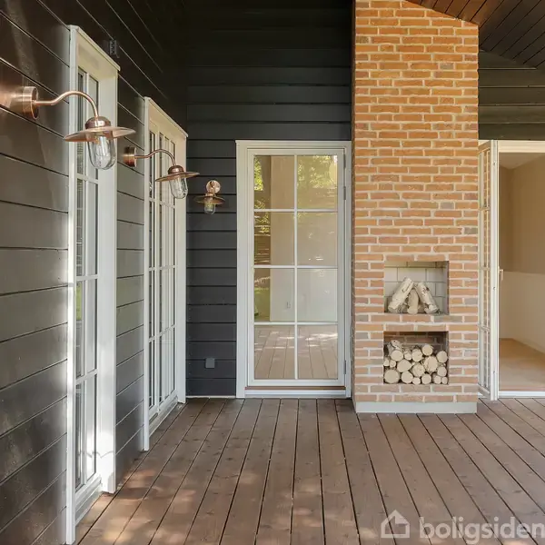 Dør med glasparti står ved siden af hvidmalet trævæg og murstenspejs, fyldt med brænde i nisjer. Udendørs står lamper, omgivet af terrasse i træ og meget naturligt lys.